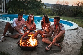 Four people sitting beside a pool roasting marshmallows around a fire.